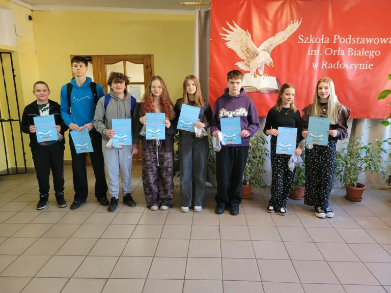 Grupa uczniów stoi w szkolnym holu przed banerem szkoły i prezentuje otrzymane dyplomy oraz nagrody po konkursie.