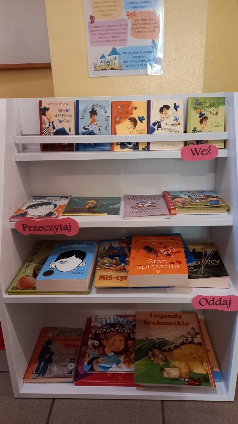 Regał z książkami w kąciku czytelniczym z oznaczeniami ‘Weź’, ‘Przeczytaj’ i ‘Oddaj’.
