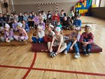 „Uczniowie siedzą na materacach w sali gimnastycznej, oglądając wydarzenie i nosząc papierowe opaski na głowach.”