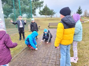 Dzieci na świeżym powietrzu mierzą taśmą odległość na chodniku, podczas gdy inni obserwują ich pracę.”