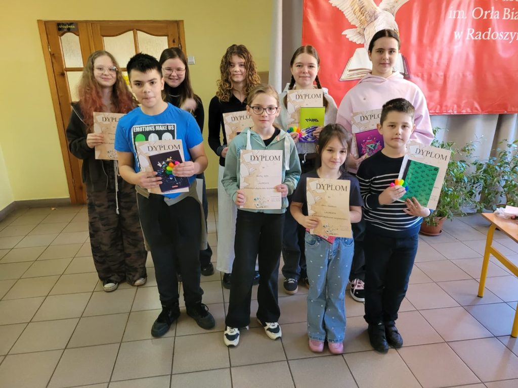 Sześcioro uczniów stoi w klasie przed ekranem z napisem o szkolnym konkursie recytatorskim i prezentuje otrzymane dyplomy oraz nagrody.