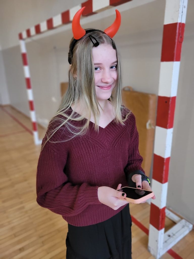 Dziewczynka z opaską z czerwonymi rogami trzyma telefon i uśmiecha się w sali gimnastycznej.