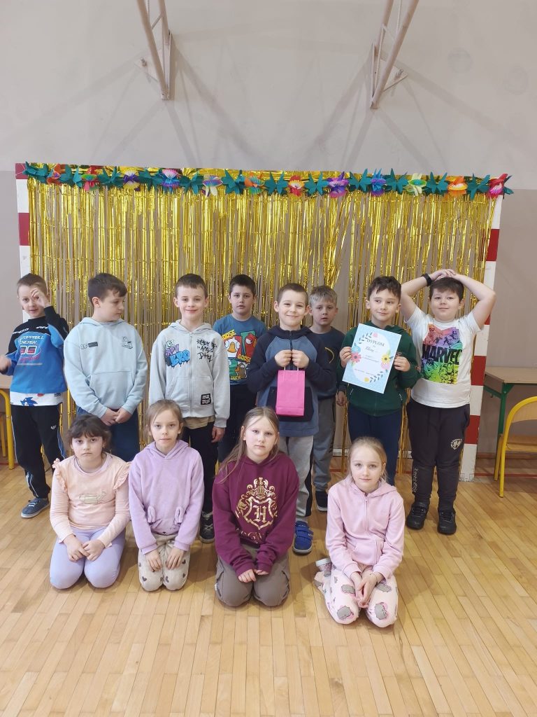 Dzieci z młodszych klas pozują w sali gimnastycznej na tle złotej dekoracji, jedno trzyma dyplom, a inne prezent.
