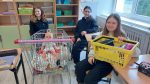 Dziewczynki siedzą w klasie obok wózka sklepowego z lalką i koszyka z przyborami, prezentując nietypowe zamienniki plecaków