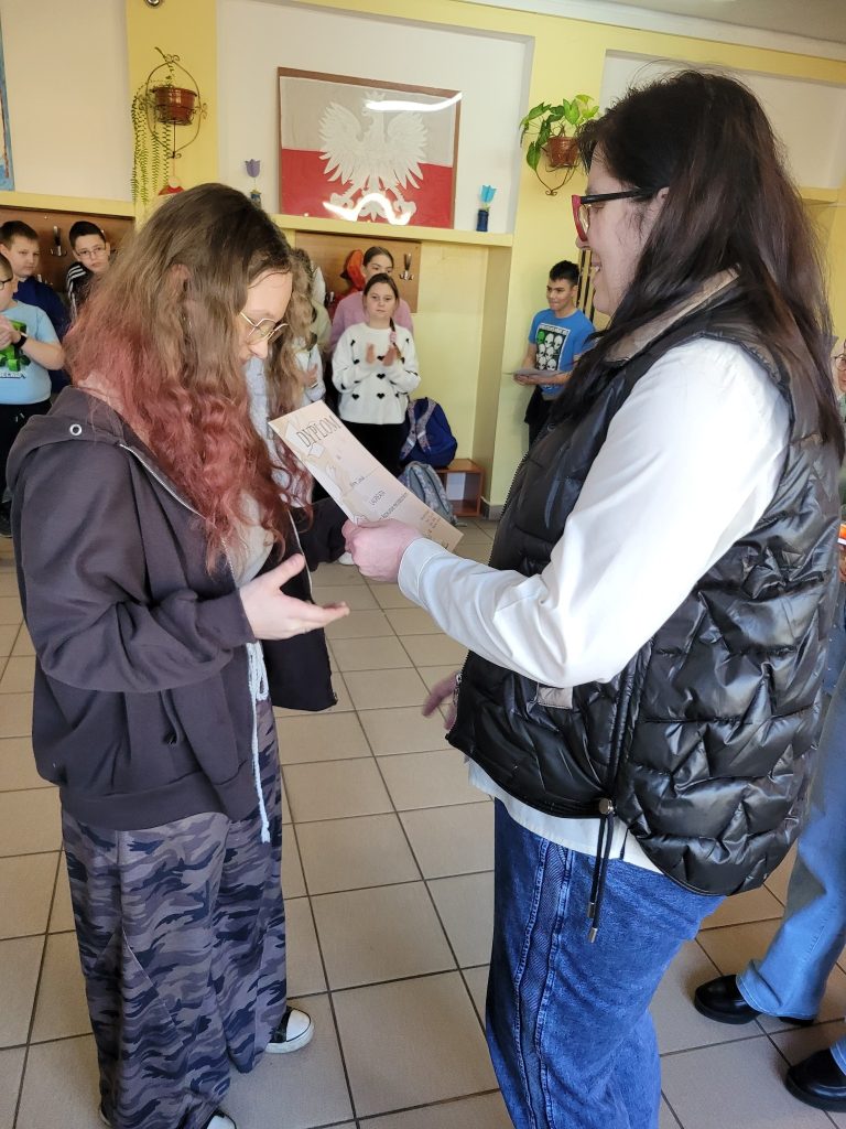Uczennica odbiera dyplom podczas szkolnego wręczenia nagród, a inni uczniowie stoją i obserwują wydarzenie w tle.