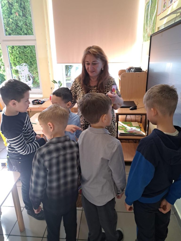 Nauczycielka w klasie rozmawia z grupą chłopców, którzy przynieśli kartki i kwiaty z okazji Dnia Kobiet.