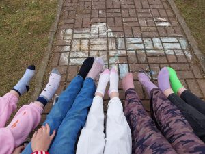 Uczniowie siedzą na chodniku z wyciągniętymi nogami, prezentując kolorowe, różne skarpetki.