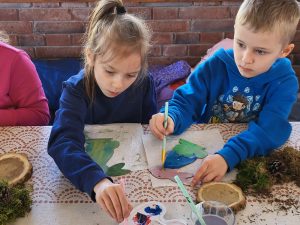 Dwoje dzieci malujących farbami drewniane figurki podczas warsztatów.