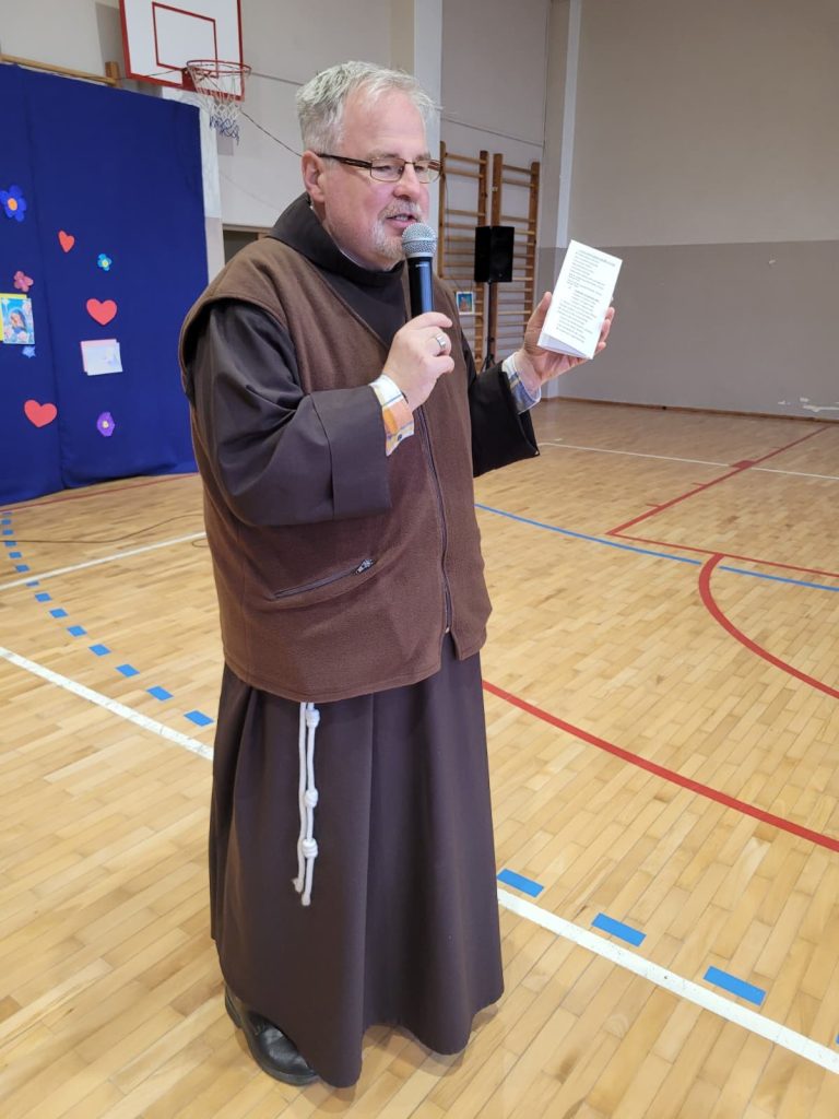 Duchowny przemawia do uczniów w sali gimnastycznej, trzymając mikrofon i kartkę z tekstem.