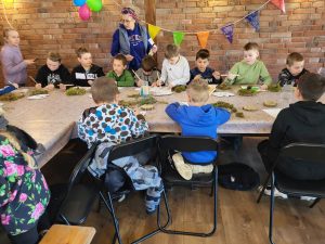 Grupa dzieci podczas warsztatów plastycznych przy stole pod okiem opiekuna.