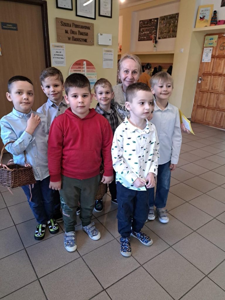 Grupa chłopców stoi z nauczycielką w szkolnym korytarzu, przygotowana do wręczania upominków z okazji Dnia Kobiet.