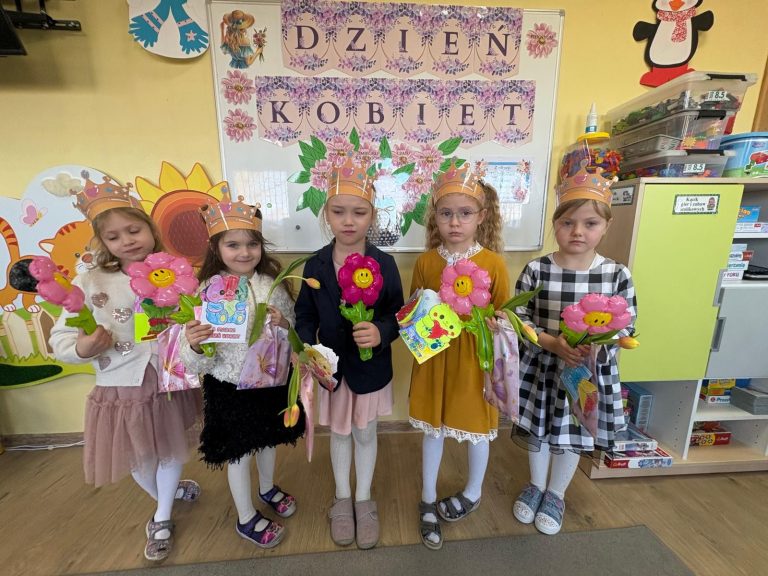 Dziewczynki w koronach stoją w klasie z kwiatami, kartkami i prezentami na tle dekoracji z napisem „Dzień Kobiet”.