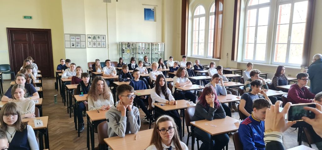 Grupa uczniów w klasie podczas konkursu języka angielskiego, siedzących przy ławkach z numerami i przygotowanych do pracy.