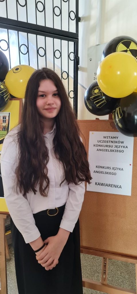 Uczennica stojąca przy tablicy ogłoszeń podczas konkursu języka angielskiego, obok dekoracji z czarno-żółtych balonów i plakatu szkolnego.