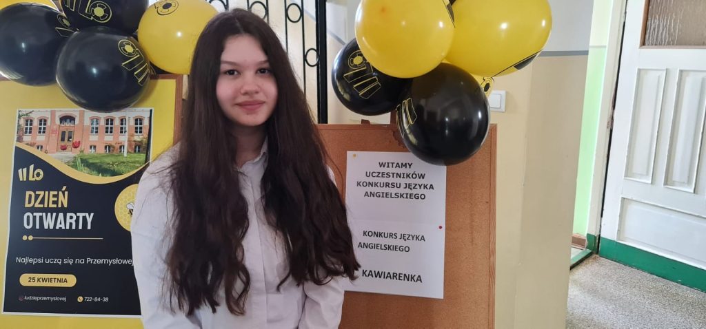 Uczennica w białej koszuli stojąca obok tablicy z informacją o konkursie języka angielskiego, w tle balony i szkolne wnętrze.
