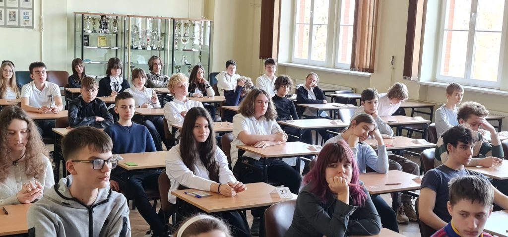 Sala lekcyjna pełna uczniów siedzących w ławkach podczas konkursu języka angielskiego, wszyscy skupieni na zadaniach.