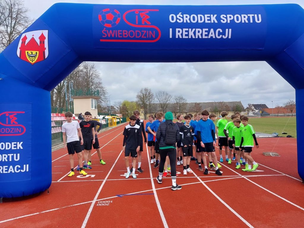 Młodzież stoi na linii startu bieżni lekkoatletycznej pod dmuchaną bramą OSiR Świebodzin przed rozpoczęciem biegu.
