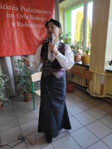 Kobieta śpiewająca i gestykulująca ręką podczas występu muzycznego w szkolnej sali.”