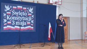 Pani dyrektor przemawia do publiczności na tle dekoracji związanej z Konstytucją 3 maja, stojąc obok polskiej flagi.