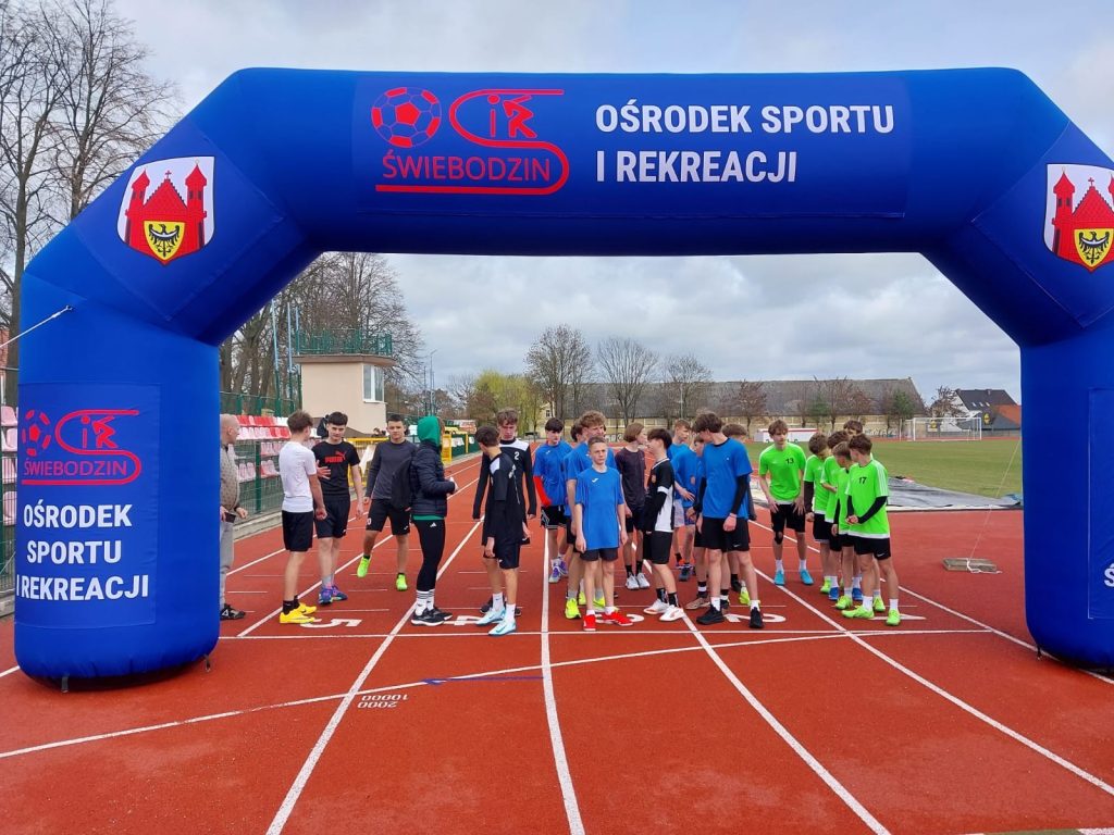 Grupa młodych zawodników przygotowuje się do startu biegu na stadionie pod bramą Ośrodka Sportu i Rekreacji w Świebodzinie.