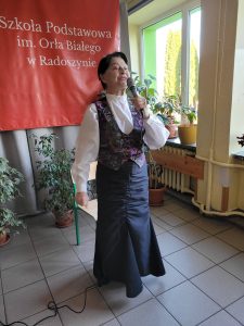Kobieta śpiewająca do mikrofonu podczas występu w szkole, stojąca przy oknie i roślinach doniczkowych.