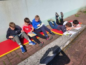 Chłopcy leżą i siedzą na matach na zewnątrz budynku, czytając książki na świeżym powietrzu.