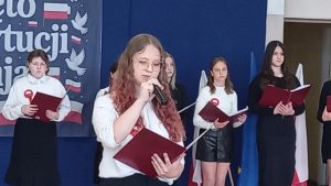 Dwie uczennice recytują do mikrofonów, trzymając czerwone teczki, podczas szkolnej akademii.