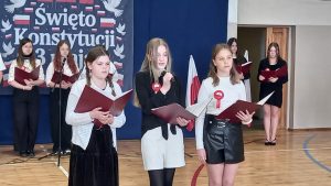 Zbliżenie na uczennice śpiewające do mikrofonu i czytające z czerwonych teczek podczas występu.