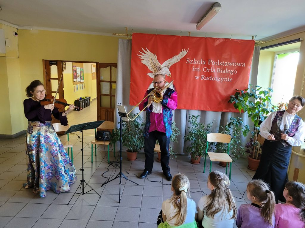 Występ muzyczny w szkole – skrzypaczka, puzonista i wokalistka występują przed siedzącymi dziećmi na tle czerwonego baneru szkoły.