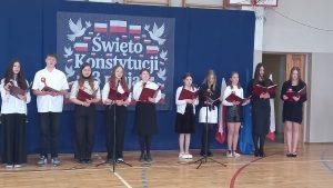 Uczniowie ustawieni w rzędzie śpiewają z nut, stojąc przed dekoracją patriotyczną i flagami.