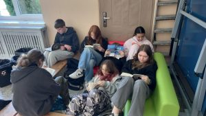 Grupa uczniów siedzi w korytarzu szkolnym na ławkach i czyta książki w swobodnej atmosferze.