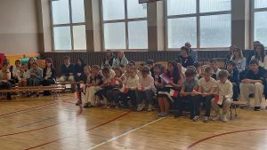 Uczniowie i rodzice siedzą w sali gimnastycznej, trzymając małe polskie flagi i obserwując występ.