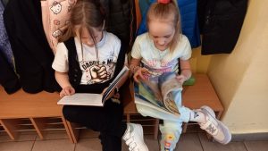 Dwie dziewczynki siedzą na ławce i czytają książki, trzymając je na kolanach.