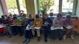 Grupa dzieci siedzących na ławce przy oknie w szkole i czytających różne książki.