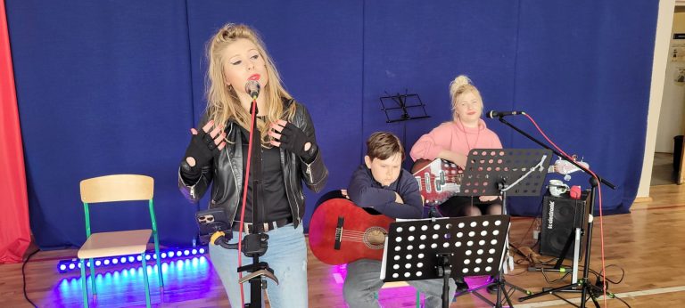 Wokalistka śpiewa do mikrofonu przy akompaniamencie młodych muzyków grających na gitarach.