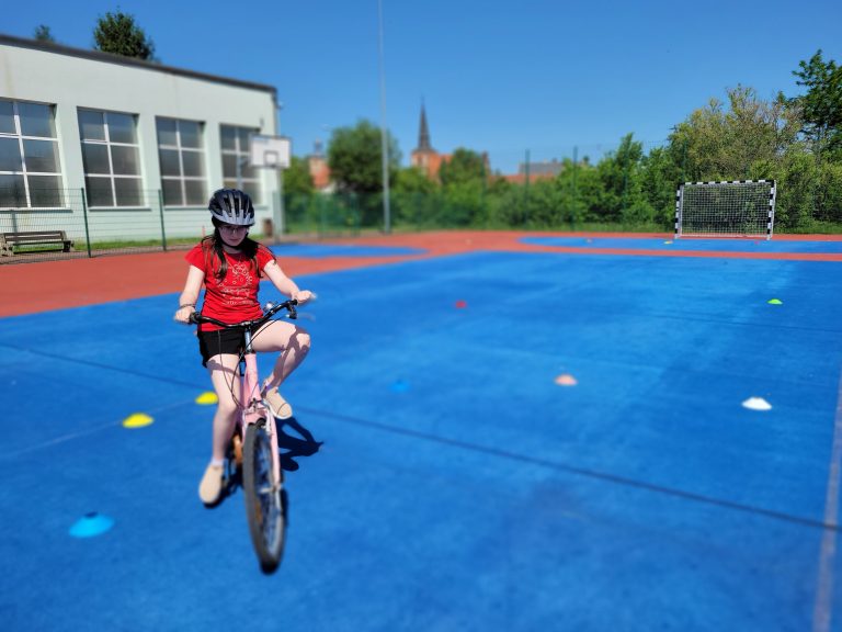 Dziewczynka na rowerze pokonuje slalom między pachołkami na szkolnym boisku.