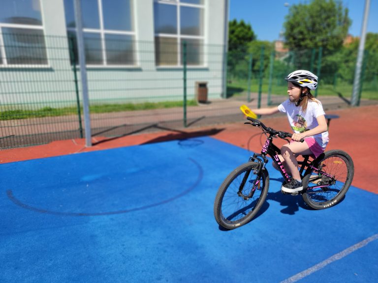 Uczennica w kasku wykonuje skręt rowerem na niebieskiej części boiska sportowego.