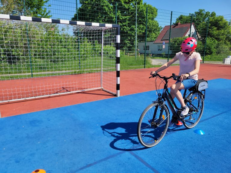 Uczennica w czerwonym kasku zatrzymała rower obok bramki sportowej na boisku.