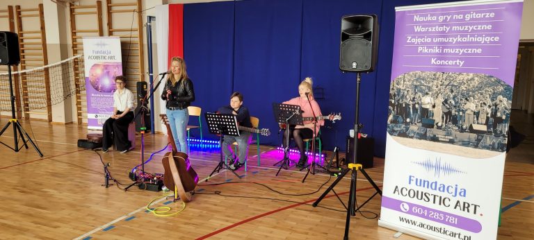 Zespół muzyczny Fundacji Acoustic Art występuje na tle scenicznej dekoracji w sali gimnastycznej.
