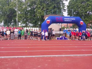 Uczniowie z różnych szkół stoją na stadionie podczas otwarcia powiatowych zawodów lekkoatletycznych.