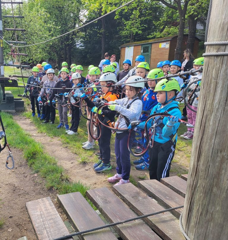 Dzieci w kaskach i uprzężach czekają w kolejce, trzymając karabińczyki przy wejściu na trasę linową.
