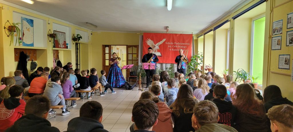 Uczniowie zgromadzeni w szkolnym holu słuchają koncertu muzyków prezentujących utwory na instrumentach dętych.
