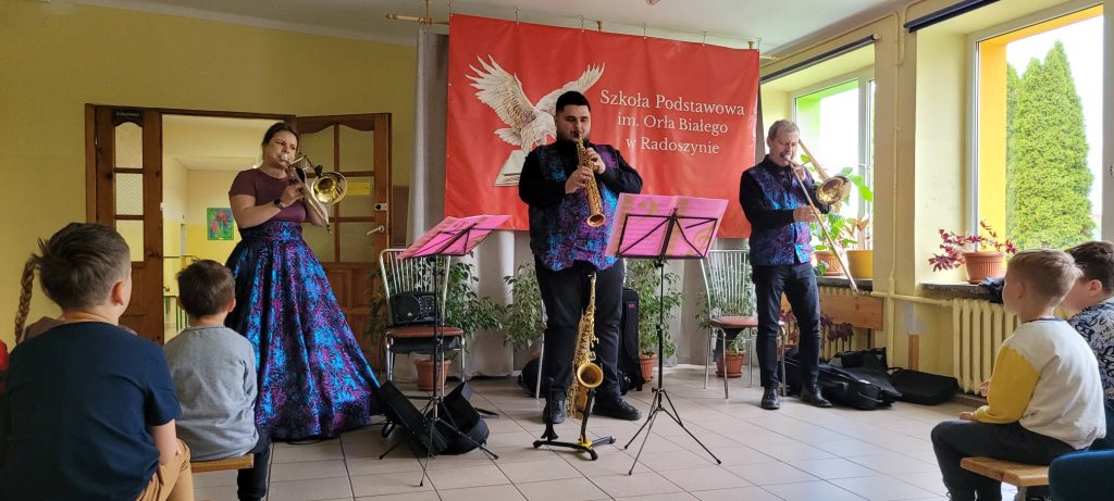 Muzycy grający na instrumentach dętych występują przed uczniami w holu Szkoły Podstawowej im. Orła Białego w Radoszynie.
