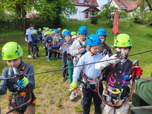 Duża grupa dzieci w kaskach i uprzężach ustawia się w kolejce przed startem na trasę linową.