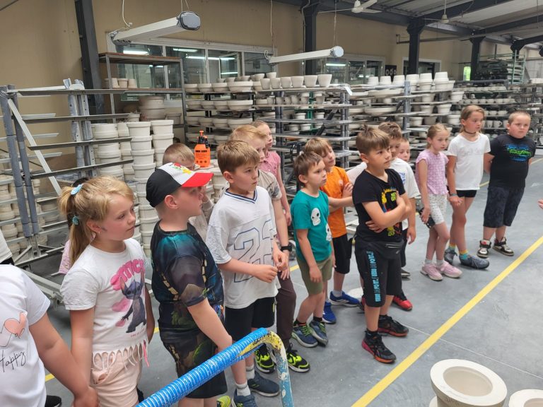 Dzieci zwiedzają halę produkcyjną pełną ceramicznych form i półek z naczyniami.