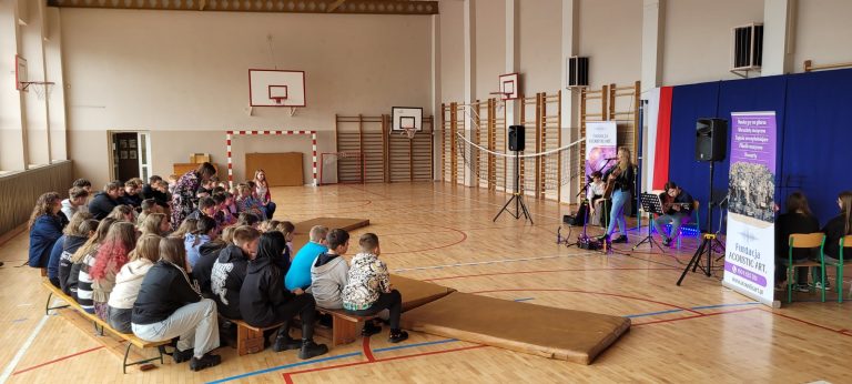 Uczniowie siedzący na ławkach w sali gimnastycznej słuchają koncertu i prelekcji Fundacji Acoustic Art dotyczącej projektu „STOP HEJT”.