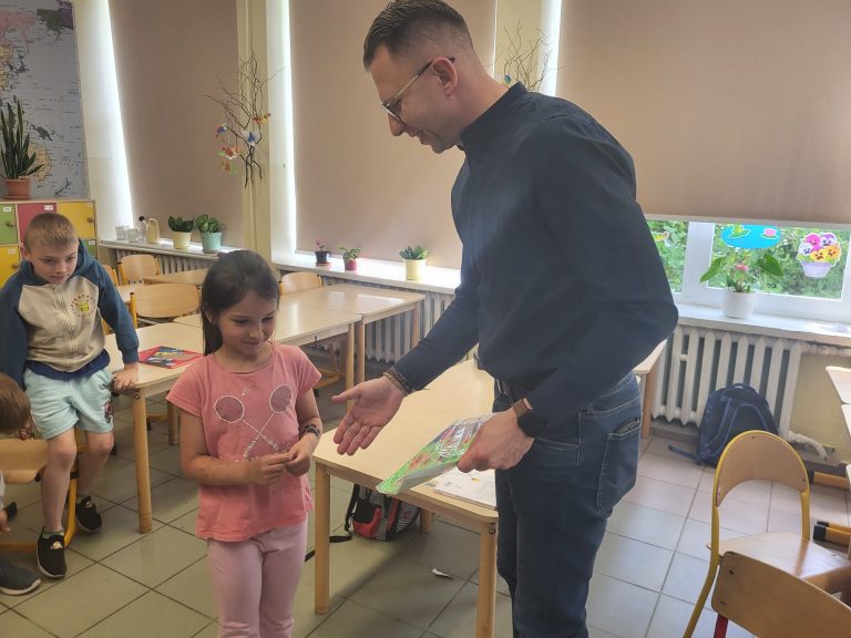 Mężczyzna wręcza uczennicy upominek lub dyplom podczas szkolnego spotkania czytelniczego.