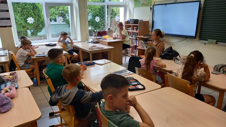 Dzieci siedzą w szkolnej klasie i słuchają głośnego czytania prowadzonego przez jedną z mam.