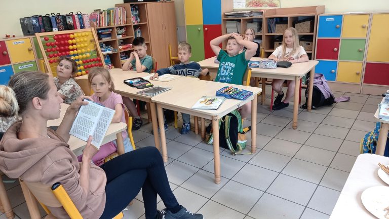Mama czyta książkę kolegom i koleżankom siedzącym w ławkach podczas zajęć promujących czytelnictwo.