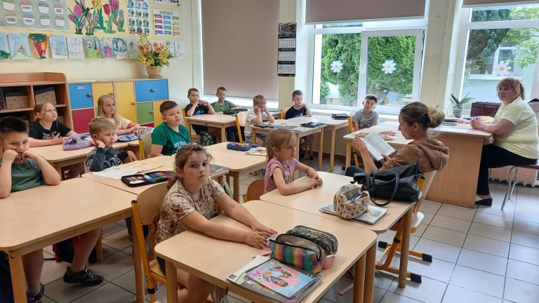 Mama czyta fragment książki dzieciom siedzącym w szkolnej klasie, a pozostali uczniowie słuchają z uwagą.